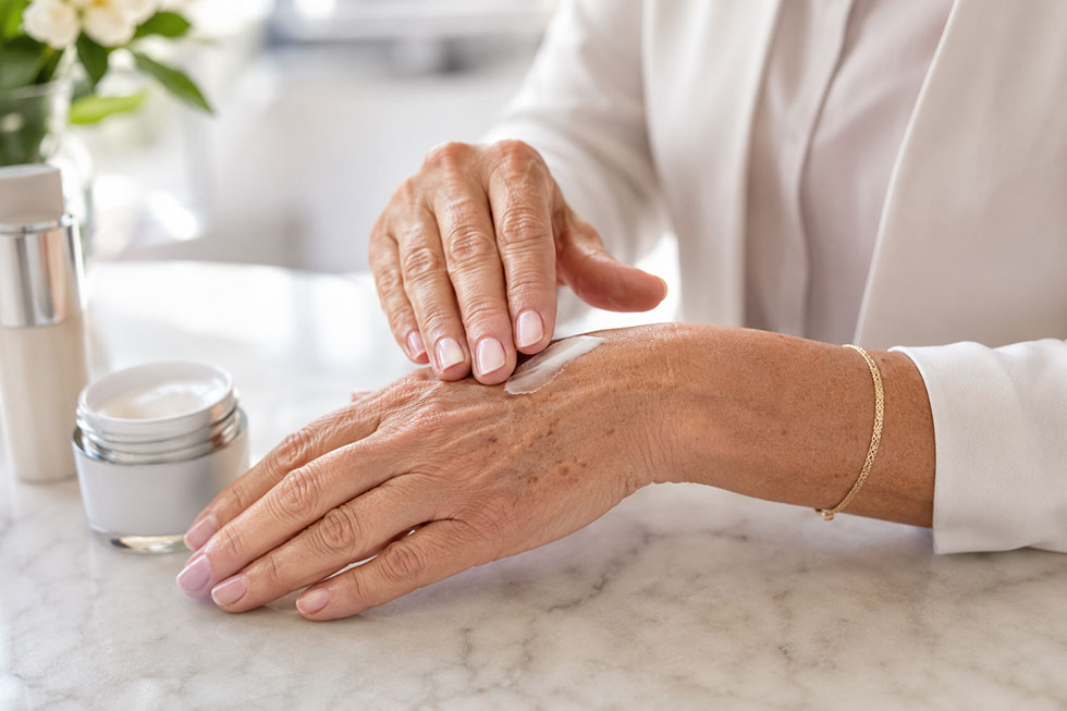 Woman applying hand cream to sun-damaged hands with dark spots and dry skin as part of a dermatologist-recommended skin care routine.